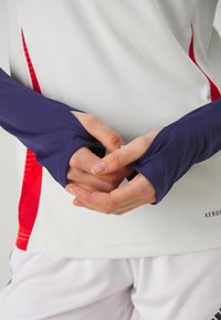 Camiseta deportiva de manga larga en blanco con acentos rojos, con mangas ajustadas de color púrpura oscuro. Textura suave, material elástico, diseño sin costuras.