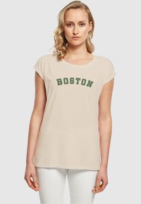 Šviesiai ruda medvilninė marškin�ė su suvyniotomis trumposiomis rankovėmis, ant krūtinės išsiuvinėtas tamsiai žalias užrašas "BOSTON". Dėvimi su baltais džinsais.