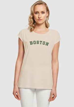 T-shirt en coton beige avec des manches courtes retroussées, présentant "BOSTON" en lettrage brodé vert foncé sur la poitrine. Porté avec un pantalon blanc.