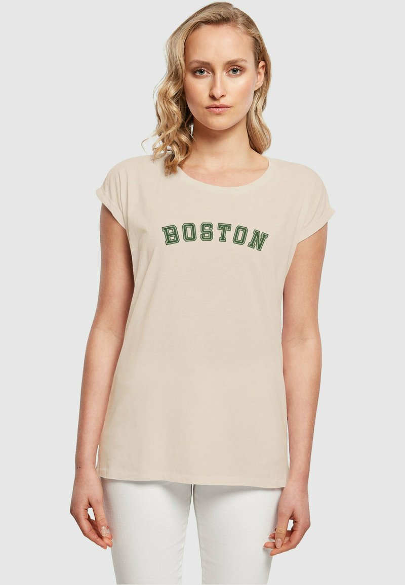 Šviesiai ruda medvilninė marškinė su suvyniotomis trumposiomis rankovėmis, ant krūtinės išsiuvinėtas tamsiai žalias užrašas "BOSTON". Dėvimi su baltais džinsais.
