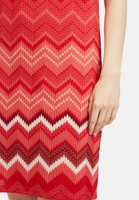 Czerwona sukienka w zygzakowy wzór z różnymi odcieniami czerwieni, bieli i czerni. Teksturowana dzianina z dopasowanym krojem; widoczna ręka.