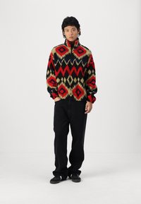 Fleece pullover in red, black, and cream with geometric patterns, high collar, and elastic cuffs. Paired with black wide-leg jeans.