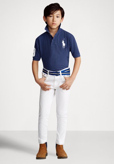 Tumši zilas polo krekls ar īsām piedurknēm un paceltu apkakli, uz kura redzams balts zirga logo. Baltas pieguļošas bikses un brūni potītes zābaki ar zilu akcenta jostu.