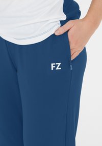 En persons hand i en marinblå byxficka med vitt "FZ"-logotyp, iklädd en vit och blå skjorta, visad från midjan till låret.
