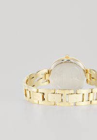 Gold-toned watch with a linked band, featuring a round case and smooth metallic finish. Engraved "GUESS" on a link and a polished back.