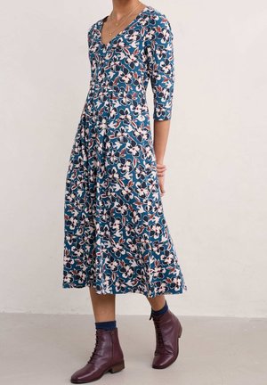 Mujer con vestido midi azul con estampado floral, mangas de tres cuartos y botines burdeos con cordones, de pie frente a un fondo liso.