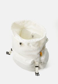 Mochila de tela blanca con una textura esponjosa, cierre en la parte superior con cordón, detalles en hardware negro y un bolsillo lateral con una etiqueta naranja brillante.