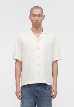 Άνδρας με ξυρισμένο κεφάλι που φοράει ένα κρεμ πουκάμισο με κοντά μανίκια και μπλε τζιν, στέκεται μπροστά σε μια απλή λευκή επιφάνεια.