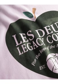 Λευκό ύφασμα με γραφικό πράσινου μήλου, που έχει κείμενο: "LES DEUX LEGACY CO." και "Για την αγάπη του παιχνιδιού." Το κείμενο είναι επικάλυψη σε έντονη, μοντέρνα γραμματοσειρά.