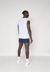 Camisola atlética branca sem mangas com um logótipo subtil nas costas, combinada com calções azul-marinho. O modelo usa sapatilhas brancas e meias até ao tornozelo.