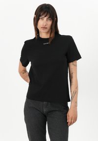 Čierne bavlnené tričko s krátkymi rukávmi, klasickým výstrihom a malým bielym logom Calvin Klein na prednej strane. Jednoduchý dizajn a rovný strih.