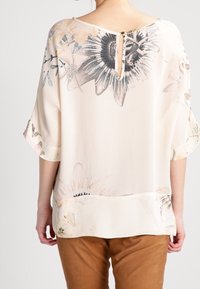 Blouse fluide couleur crème avec imprimé floral, incluant des tournesols, détail dos en keyhole et manches cloche 3/4. Tissu lisse et léger.