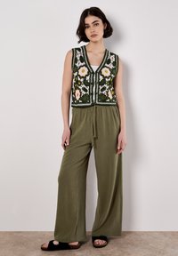 Gilet senza maniche verde con ricami floreali in giallo, bianco e arancione; abbinato a pantaloni larghi verde oliva e sandali neri con zeppa.