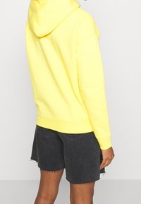 Felpa gialla con cappuccio, dal texture morbido, dotata di tasca a kangaroo e polsini a costine. Abbinata a pantaloncini di jeans neri con orlo sfrangiato.