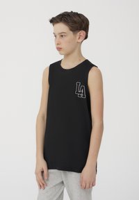 Canottiera nera senza maniche in tessuto morbido. Presenta un colletto tondo e un logo grigio "LA" sul petto. Abbinata a pantaloni grigi.