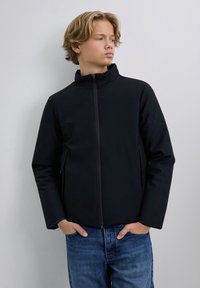 Junge Person mit blonden Haaren, die eine schwarze Zip-Jacke und blaue Jeans trägt, steht mit den Händen in den Taschen und schaut nach rechts.