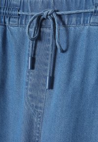 Heledad sinised teksapüksid, millel on siniste otstega nööriga vöökoht ja küljetasku, näidates sujuvat, veidi tekstuuri omavat kangast.