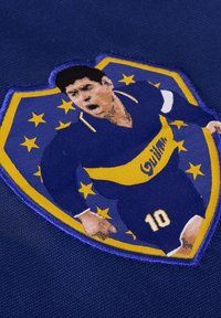 Blauwe stoffen badge met een ontwerp van een geel en blauwe voetbalshirt met een spelerfiguur, nummer 10, en sterren op de achtergrond.