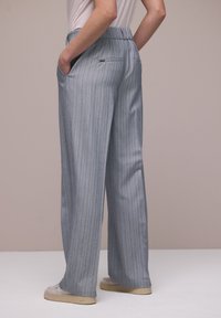 Personne portant un pantalon large taille haute à rayures grises claires, les mains dans les poches avant et des baskets beiges sur un sol blanc.