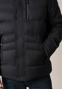 Veste noire matelassée avec fermeture éclair et bouton, dotée de deux poches avant et d'un tissu texturé. Design chaud et isolé.