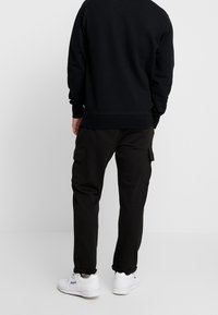 Sweat-shirt noir avec poignets côtelés, associé à un pantalon cargo foncé avec poches latérales. Porté avec des baskets blanches, mettant en valeur une tenue décontractée.
