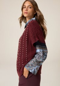 Burgundowa sweter z dzianiny z wzorem w warkocz i srebrnymi ćwiekami, nałożony na niebieską koszulę w paisley z szerokimi rękawami. Gładkie tło.