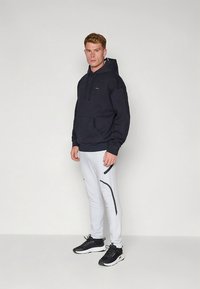 Laivastonsininen huppari etutaskulla ja kiristysnarulla, yhdistetty vaaleanharmaisiin jumppahousuihin, joissa on sivutasku ja mustat yksityiskohdat, yhdistetty mustiin lenkkareihin.