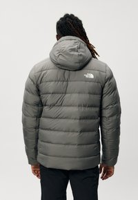 Geacă puffer gri cu design matlasat orizontal, glugă și logo pe partea superioară a spatelui. Textură netedă, potrivită pentru vreme rece.