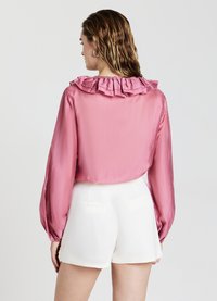 Blusa di raso rosa con colletto arricciato, maniche lunghe e polsini elasticizzati, abbinata a pantaloni corti bianchi su misura.