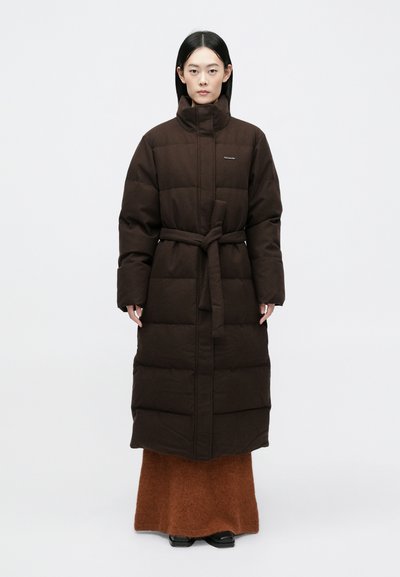 Manteau long rembourré marron avec col montant et ceinture, présentant un motif matelassé horizontal. Porté par-dessus une jupe texturée marron et des chaussures noires.