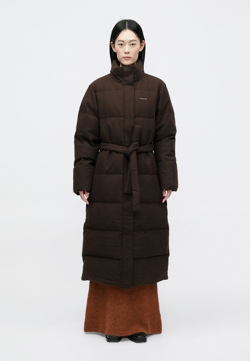 Brun polstret lang frakke med høj krave og bælte, der har et vandret quiltet mønster. Båret over en brun tekstureret skørt og sorte sko.