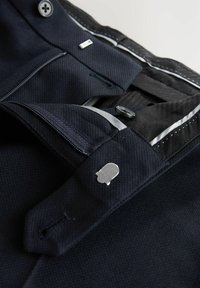 Laivastonsiniset housut, joissa teksturoitu kangas, hopeiset metalliosat, nappisulkimet ja korostetut tikkausompeleet. Sisätaskut näkyvissä.