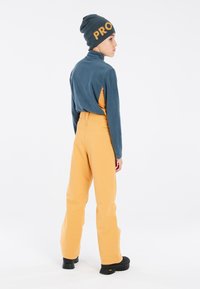Fleece coltrui in teal, met gele accenten, gecombineerd met gele high-waisted broek. Zwarte platform schoenen en een blauwe beanie met de tekst "PRO".