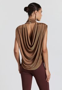 Haut drapé marron avec un col montant et un dos ouvert, présentant une texture brillante et des boutons décoratifs sur le col. Associé à des leggings marron.