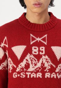 Kötött piros pulóver krém színű geometrikus mintákkal, hegyekkel és "G-Star Raw" szöveggel. Kerek nyakkivágással és bordázott textúrával rendelkezik.