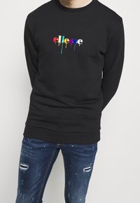 Felpa nera con un logo "ellesse" colorato e gocciolante sul davanti. Indossata con jeans blu strappati con patch strappati.
