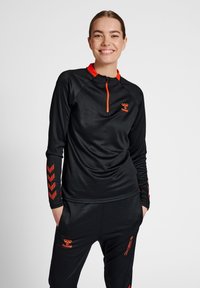 Maglietta sportiva nera a maniche lunghe con collo a zip quarti, colletto e dettagli rossi a contrasto, realizzata in tessuto texturizzato e con motivi a chevron sulle maniche.