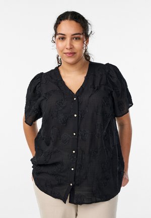 Žena s kučeravými vlasmi v čiernej textúrovanej blúzke na zapínanie a svetlých nohaviciach, stojaca pred bielym pozadím.