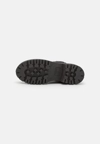 Semelle de botte noire avec des crampons profonds et décorés, comprenant des formes en croix et une section talon rectangulaire, sur fond blanc.