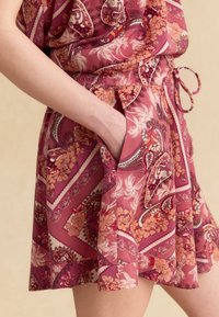 Mano descansando en el bolsillo de una falda rosa con patrón floral y paisley naranja, cintura atada y textura de tela suave.