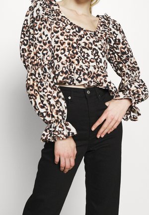 Nainen, joka on pukeutunut leopardikuvioiseen pitkähihnaiseen puseroon, jossa on röyhelöiset kalvosimet, ja mustiin korkeavyötäröisiin housuihin, poseeraa yhdellä kädellä lantiolla.