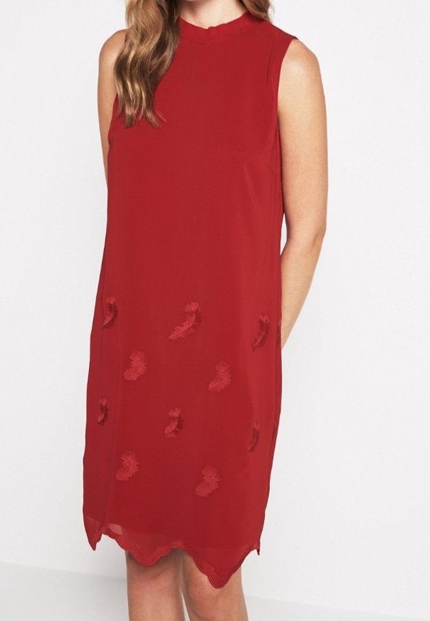 Robe rouge sans manches avec col montant, détails floraux texturés et ourlet festonné portée par une femme aux cheveux châtain clair ondulés.