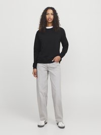 Vrouw met krullend haar die een zwarte sweatshirt, lichtgrijze jeans en witte sneakers draagt, staand met één hand in de zak op een effen achtergrond.