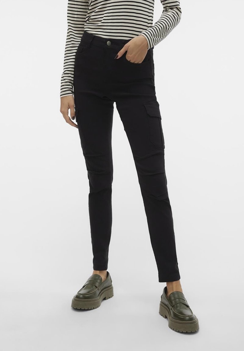 Personne portant un pantalon cargo noir, une chemise à rayures à manches longues et des mocassins épais vert olive, debout sur un fond blanc.