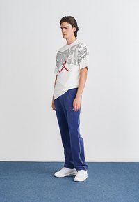 Fehér póló fekete grafikai dizájnnal és piros logóval, kék melegítőnadrággal, amelynek fehér oldalsávjai vannak, valamint fehér sportcipővel.