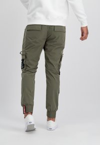 Mann in olivgrünen Cargo-Hosen mit elastischen Bündchen und einem weißen Sweatshirt, der vor einem schlichten hellen Hintergrund steht.