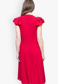 Punainen mekko, jossa on lyhyet röyhelösleeveet ja kiinnitetty vartalo, joka leimahtaa polveen asti ulottuvaan hameeseen. Sujuva kangas, jossa on yhtenäinen väri.