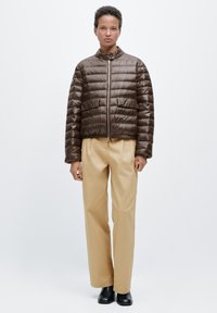 Veste matelassée marron avec coutures horizontales, fermeture éclair devant et col montant. Associée à un pantalon large crème et des bottines noires.
