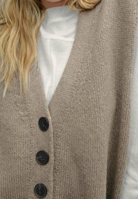 Vesti virkattu vaaleanbeige, jossa on syvä V-pääntie ja neljä mustaa nappia edessä sekä teksturoitu kangas. Päällä valkoinen pitkähiha-toppi.