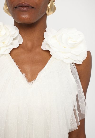 Witte jurk met textuurrimpels, voorzien van verhoogde witte bloemenaccenten op de schouders en een V-halslijn. Doorzichtig stof met stippen.